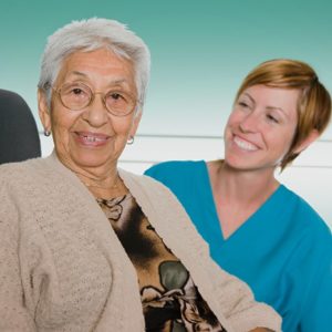 An elderly woman smiling at the camera in a seated position while a healthcare professional looks at them and smiles while positioned behind them.
