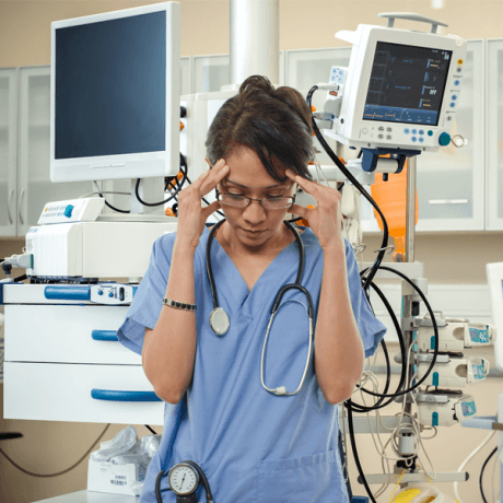 A nurse standing with hands placed on her temples in pain as medical monitoring systems are in the background.