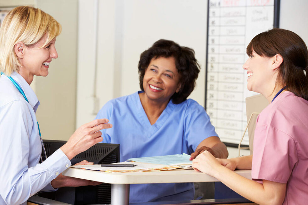 Three healthcare professionals, two wearing scrubs and one in a lab coat, engaging in a discussion at a nurse's station.