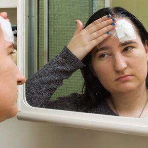 A woman examines her reflection in a bathroom mirror, touching a bandage over her left eyebrow with a concerned expression.