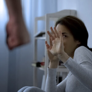A woman in a domestic setting appears fearful, holding up her hands defensively against an unseen aggressor.