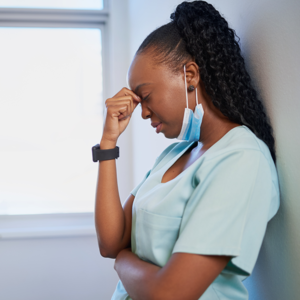 A healthcare worker, a Black woman with long braided hair, appears stressed, resting her forehead on her hand while standing against a light-colored wall next to a window.