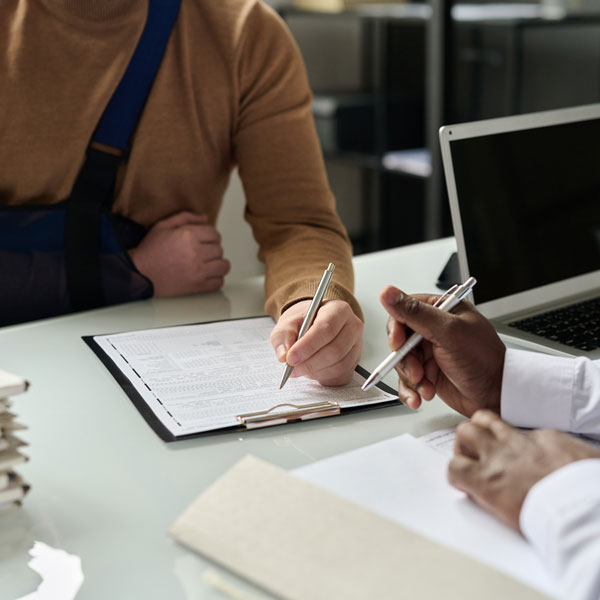 Two people are sitting at a desk, one with a pen poised over a form on a clipboard, and the other holding a pen. One person is wearing a beige sweater and has their arm in a sling, while the other person is wearing a white shirt. A laptop is open on the desk.