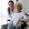 Nursing_Home_Admin_NHA A young female healthcare professional stands behind an older woman sitting in a wheelchair, both smiling at the camera.