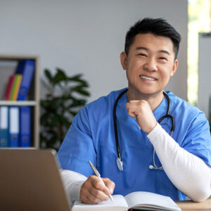 A smiling healthcare professional in blue scrubs, wearing a stethoscope around their neck, is sitting at a desk with a laptop and an open notebook. The individual appears to be working or taking notes. The background shows binders and a plant, suggesting a work environment, possibly a clinic or office.