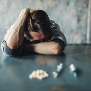 A person with a distressed expression rests their head on their arm on a table. In front of them, there are scattered pills and syringes, symbolizing substance abuse or addiction.