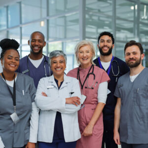 A diverse group of six healthcare professionals smiling and standing together in a modern medical facility. They are wearing scrubs and lab coats, with stethoscopes around their necks, representing teamwork and professionalism in healthcare.
