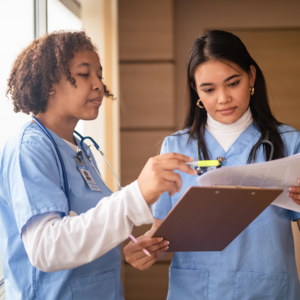 Two healthcare professionals in light blue scrubs reviewing a clipboard together in a well-lit indoor setting. One holds the clipboard and a highlighter, while the other points to the document. Both appear focused on their task.