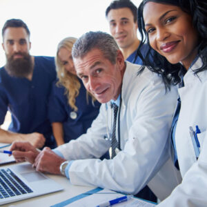 A group of healthcare professionals, including doctors and nurses, gathered around a laptop, smiling and engaged in discussion.