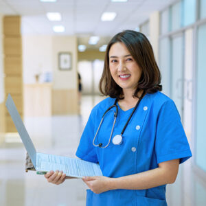 Smiling female nurse in blue scrubs holds a patient chart in a well-lit hospital hallway.