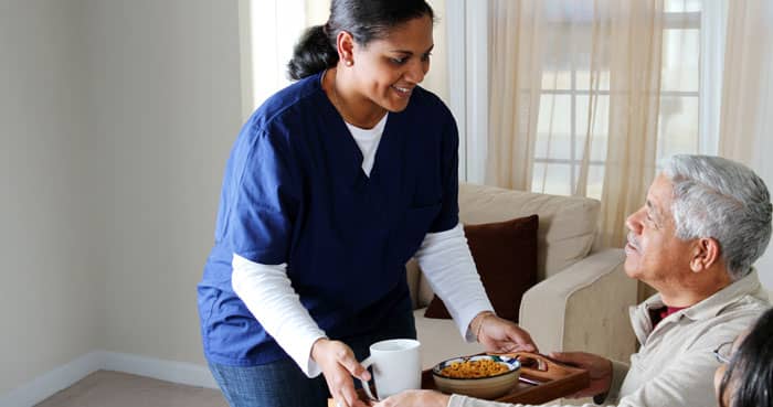 Home care aide in blue scrubs smiling while serving breakfast on a tray to an older adult man seated on a couch in a home setting