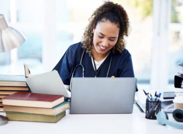 Smiling nurse completing self-paced continuing education online at a laptop beside books and work materials.