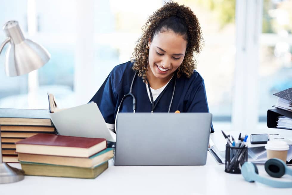 Smiling nurse completing self-paced continuing education online at a laptop beside books and work materials.