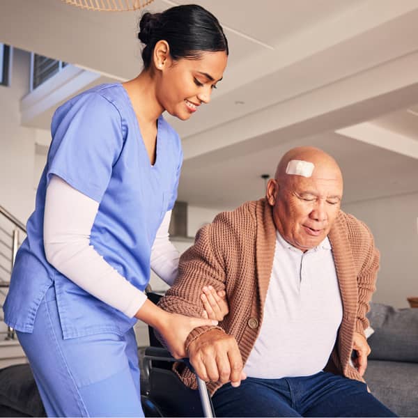 Healthcare caregiver assisting an older adult with a head bandage from a wheelchair in a residential care setting.